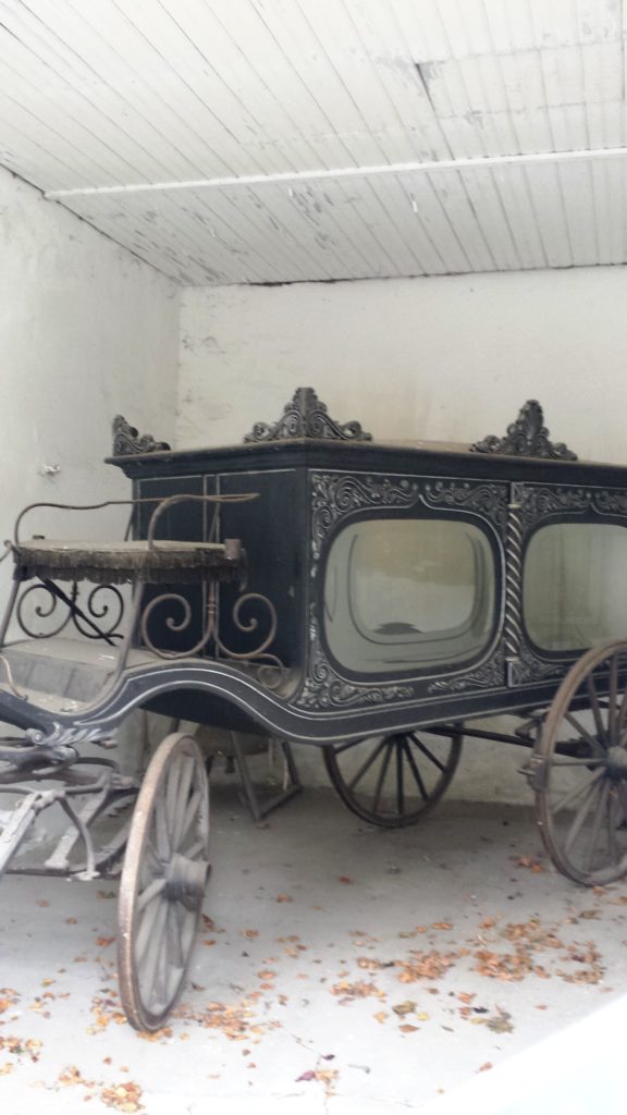 Gegenüber der katholischen Kirche kannst du einen alten Leichenwagen betrachten